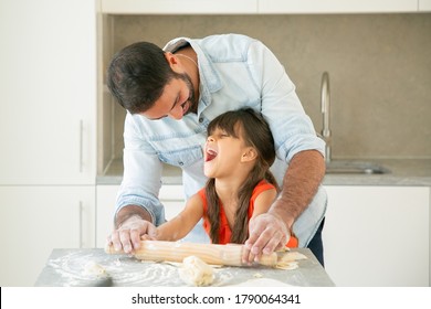 Excited Girl And Her Happy Dad Rolling And Kneading Dough On Kitchen Table With Flour Messy. Father Teaching Daughter To Bake Bread Or Pies. Family Cooking Concept