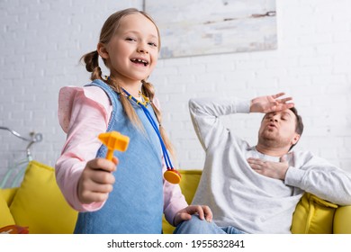 Excited Girl Holding Toy Reflex Hammer While Playing Doctor Near Dad Pretending Sick On Blurred Background