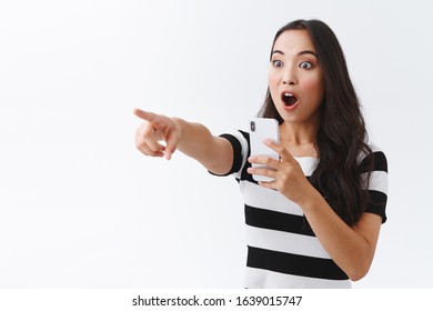 Excited girl witness something extremely awesome, record video on smartphone camera, pointing and staring impressed sideways, gasping, drop jaw astonished, standing white background
