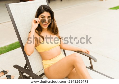 Excited gorgeous woman in a bikini putting on sunglasses while relaxing in the pool lounger feeling happy