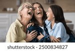 Excited grandma, happy young mother and cheerful kid girl laughing out loud with open mouth, having fun, telling jokes on home couch, tickling each other, enjoying family leisure. Banner shot