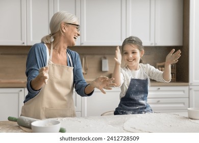 Excited grandmother and happy little granddaughter girl in aprons throwing flour over table with ingredients laughing, having fun, clapping floury hands for making cloud, playing while cooking - Powered by Shutterstock