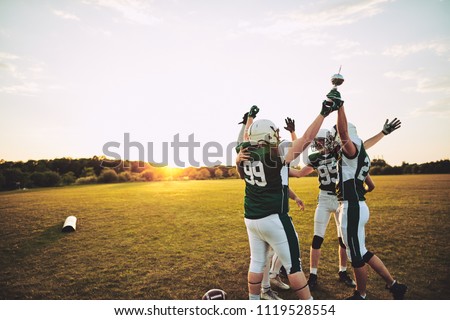 Excited group of American football players standing together in a huddle and raising a championship trophy in celebration