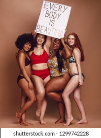 Excited group of women holding a every body is beautiful signboard over brown background. Multi-ethnic females of different figure and size looking excited together with a placard.