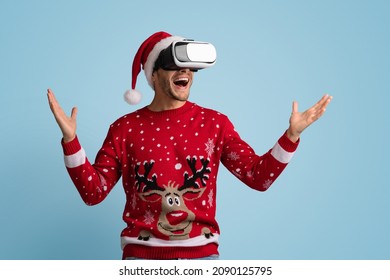 Excited Guy Wearing Vr Glasses Headset And Christmas Sweater Experiencing Virtual Reality While Standing On Blue Studio Background, Emotional Man In Santa Hat And Goggles Enjoying Virtual Xmas Party