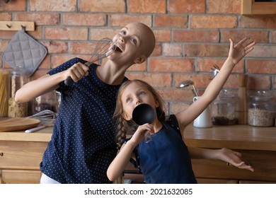 Excited Hairless Cancer Mom And Funny Little Daughter Girl Playing Concert Show, Singing At Kitchen Utensils While Cooking, Listening To Music, Songs On Radio, Having Fun, Enjoying Leisure At Home