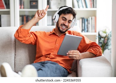 Excited handsome bearded middle eastern young man relaxing on couch at home, using brand new digital tablet and stereo wireless headphones, having online paty with friends, watching video, copy space