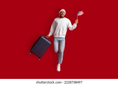 Excited handsome millennial african american guy in casual and Santa hat holding American flag, carrying suitcase, jumping over red studio background, travelling to the US on xmas holidays, copy space