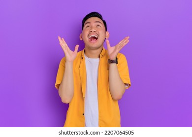 Excited Handsome Young Asian Man Looking Up With A Happy Expression While Raising Arms Isolated On Purple Background