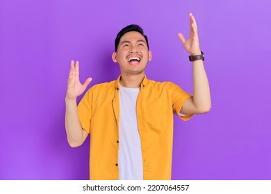 Excited Handsome Young Asian Man Looking Up With A Happy Expression While Raising Arms Isolated On Purple Background