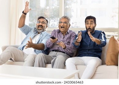 Excited happy adult Indian men of three family generations watching sport match on TV, celebrating team winning, laughing, shouting, waving hands, using remote control device, sitting on couch - Powered by Shutterstock