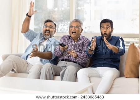 Excited happy adult Indian men of three family generations watching sport match on TV, celebrating team winning, laughing, shouting, waving hands, using remote control device, sitting on couch
