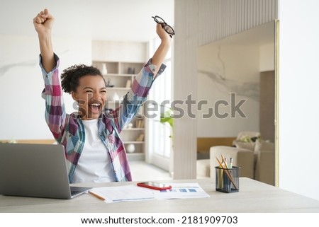 Excited happy african american girl student got good exam test scores on laptop. Overjoyed teenage schoolgirl raising hands screaming celebrates personal achievement or finishing homework.