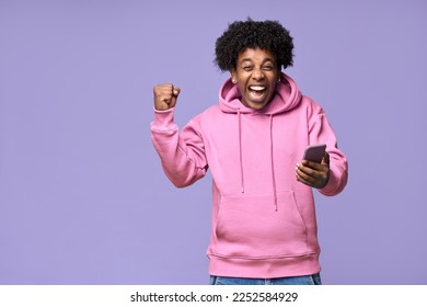 Excited happy African teen guy winner holding cell phone winning game online on cellphone, using mobile apps technology celebrating victory receiving message isolated on light purple background. - Powered by Shutterstock