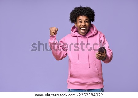 Excited happy African teen guy winner holding cell phone winning game online on cellphone, using mobile apps technology celebrating victory receiving message isolated on light purple background.