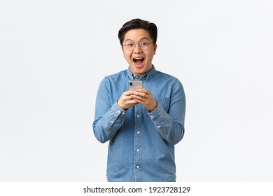 Excited Happy Asian Man Reacting Cheerful At Awesome News Read Online, Holding Mobile Phone And Looking Thrilled With Event Coming Up. Guy Download Cool New App Or Game, White Background