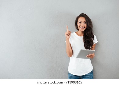 Excited happy asian woman holding tablet computer and pointing finger up at copy space while standing isolated over gray background