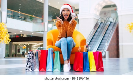 Excited Happy Beautiful Young Woman In Santa Hat With Many Colorful Bright Shopping Bags Is Sitting On The Chair In The Mall During Christmas Holiday