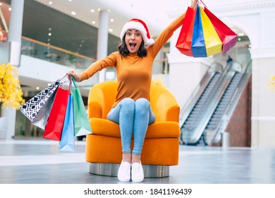 Excited Happy Beautiful Young Woman In Santa Hat With Many Colorful Bright Shopping Bags Is Sitting On The Chair In The Mall During Christmas Holiday