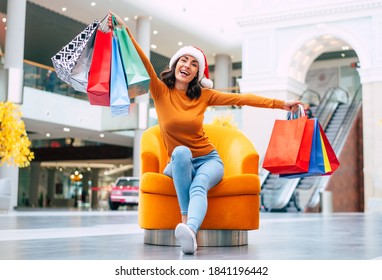 Excited Happy Beautiful Young Woman In Santa Hat With Many Colorful Bright Shopping Bags Is Sitting On The Chair In The Mall During Christmas Holiday