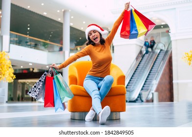 Excited Happy Beautiful Young Woman In Santa Hat With Many Colorful Bright Shopping Bags Is Sitting On The Chair In The Mall During Christmas Holiday