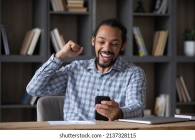 Excited happy Black guy using mobile phone at workplace table, feeling joy, getting good news, reading message on smartphone, celebrating win, success, achieve, making winner yes hand - Shutterstock ID 2137328115