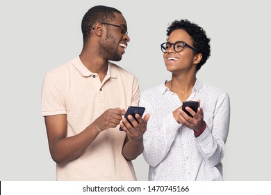 Excited happy black millenial family spouse in eyeglasses holding smartphones , looking at each other, received sms notification with big sale discount price, isolated on grey studio background.