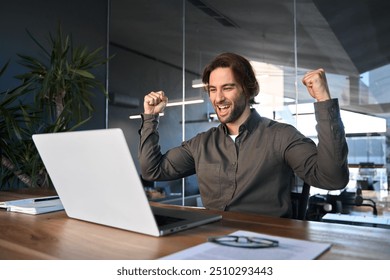 Excited happy business man winner celebrating success looking at laptop in office. Young professional businessman executive rejoicing financial growth good work results, winning corporate profit goal. - Powered by Shutterstock