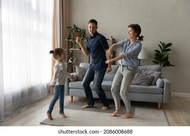 Excited Happy Couple Of Parents Teaching Cheerful Daughter Girl To Dance In Living Room. Mom, Dad And Kid Enjoying Activity At Home, Exercising, Having Fun To Music, Celebrating Moving Into New House