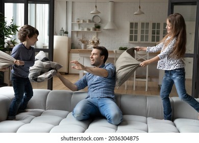 Excited Happy Dad And Crazy Sibling Kids Fighting With Pillows On Couch, Enjoying Funny Battle With Cushions. Millennial Father And Children Playing Wild Active Games At Home, Having Fun Together