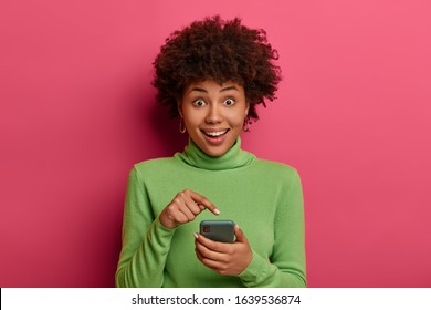 Excited happy ethnic female points at smartphone display, glad to receive romantic date invitation, wears casual green poloneck, sends new just made selfie on web page, stands in vivid studio - Shutterstock ID 1639536874
