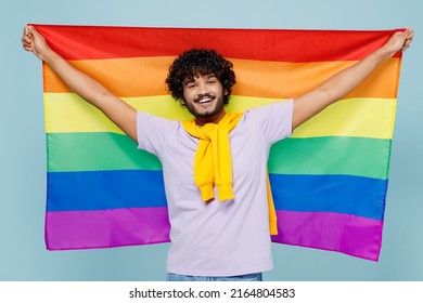 Excited happy fancy jubilant young bearded Indian man 20s years old wears white t-shirt hold rainbow colorful striped flag looking camera isolated on plain pastel light blue background studio portrait
