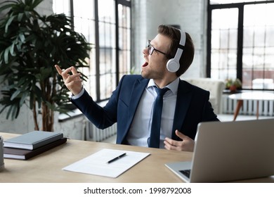Excited happy funny businessman in wireless headphones listening to music, enjoying favorite song, playing air guitar, pretending to be guitarist, rock star, relaxing and having fun during work break - Shutterstock ID 1998792008
