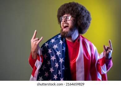 Excited happy hipster man with Afro hairstyle wrapped in american flag, rejoicing, celebrating national holiday, showing v sign. Indoor studio shot isolated on colorful neon light background.