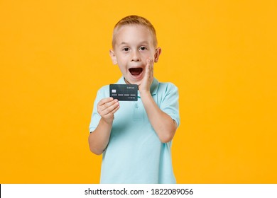 Excited happy little fun male kid boy 5-6 years old wearing stylish blue turquoise t-shirt polo hold in hand mockups of credit bank card isolated on yellow color wall background child studio portrait