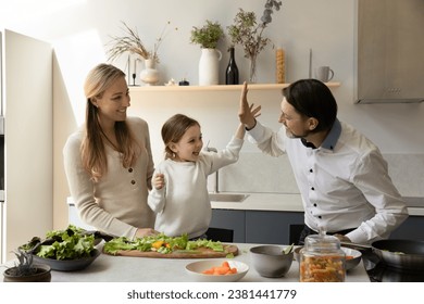 Excited happy little kid girl giving high five to chef dad, helping parents to prepare healthy meal for dinner in kitchen. Family cooking salad, having fun at table with cut vegetables - Powered by Shutterstock