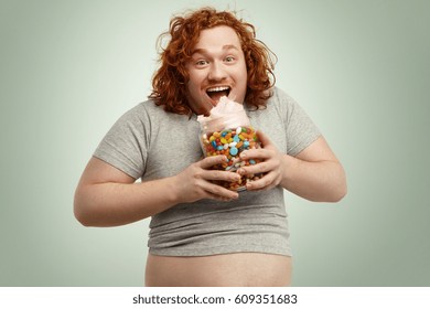 Excited Happy Obese Young Man With Ginger Curly Hair Opening Mouth Widely While Eating Candy Floss Out Of Glass Jar Full Of Colorful Sweets. Obesity, Gluttony And Unhealthy Lifestyle Concept