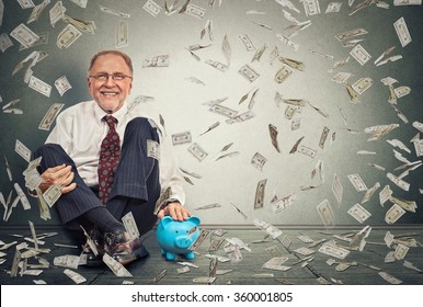 Excited happy senior man sitting on a floor with piggy bank under a money rain isolated on gray wall background. Positive emotions financial success luck good economy concept