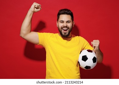 Excited happy young bearded man football fan in yellow t-shirt cheer up support favorite team hold soccer ball celebrate clenching fists say yes isolated on plain dark red background studio portrait
