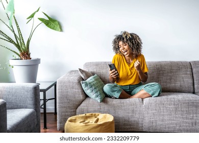 Excited happy young black woman holding smart phone device sitting on sofa at home - Happy satisfied female looking at mobile smartphone screen gesturing yes with clenched fist - Technology concept - Powered by Shutterstock