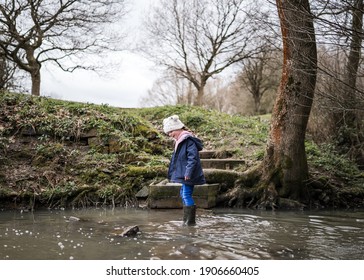 Excited Happy Young Girl Cute In Wellington Boots Walking Autumn Countryside In River Stream Splashing In Water Exploring The Great Outdoors Nature Park With Bobble Hat And Blue Coat In Winter Nature