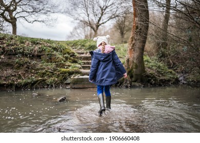 Excited Happy Young Girl In Wellington Boots Walking Through Autumn Countryside River Stream Splashing In Water Exploring The Great Outdoors Nature Park With Bobble Hat And Blue Coat In Winter Flowing