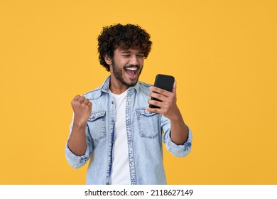 Excited happy young indian man winner feeling joy using smartphone winning lottery game, betting, getting cashback online gift in mobile app message holding cell phone isolated on yellow background. - Shutterstock ID 2118627149