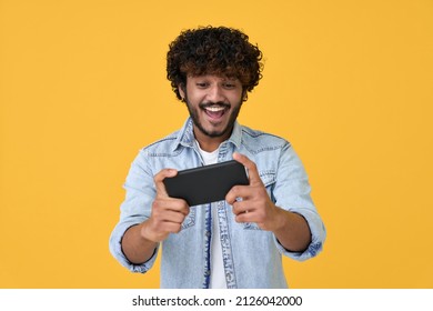 Excited Happy Young Indian Man Gamer Player Using Smartphone Playing Game In Mobile Videogame App Or Watching Digital Video Stream Holding Cell Phone Isolated On Yellow Background.