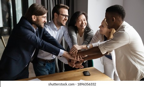 Excited happy young and mature mixed race colleagues joining hands, strengthening team spirit, motivating each other at meeting. Laughing diverse multiracial business people celebrating shared success