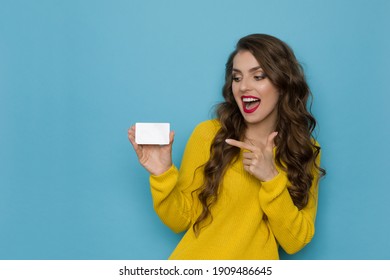 Excited And Happy Young Woman In Yellow Sweater Is Holding White Plastic Card, Pointing At It And Talking. Waist Up Studio Shot On Blue Background.