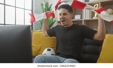 Excited hispanic man with a beard cheering at home while watching a soccer game holding a flag and sitting on a couch. - Powered by Shutterstock