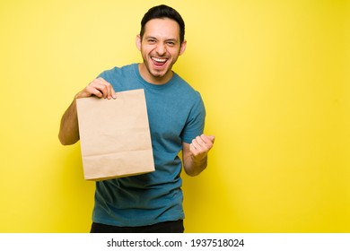 Excited Hispanic Man Happy To Receive His Takeout Food For Lunch. Latin Man Smiling And Holding A Brown Bag With Delivery Food