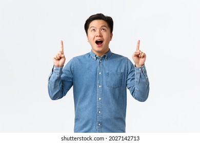 Excited And Impressed Asian Young Man Open Mouth, Saying Wow Fascinated, Checking Out Promo, Looking And Pointing Fingers Up At Advertisement, Stare At Awesome Banner, White Background