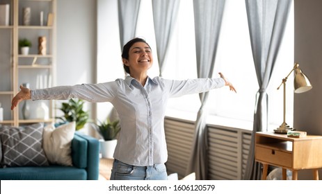 Excited independent young indian woman housewife first time home owner stand in modern living room interior with arms outstretched enjoy free lifestyle freedom wellbeing dance alone in own apartment